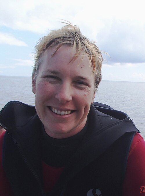 Andrea Baldwin smiles at the camera, wearing a wetsuit with the ocean behind her.