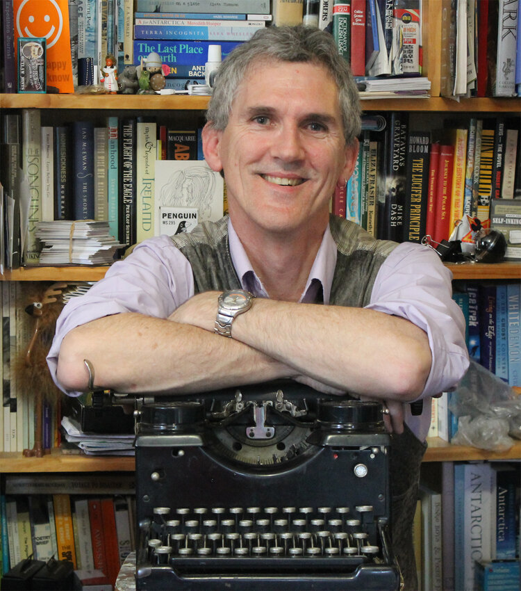 Craig leans on a typewriter, smiling. Behind him is a bookshelf.