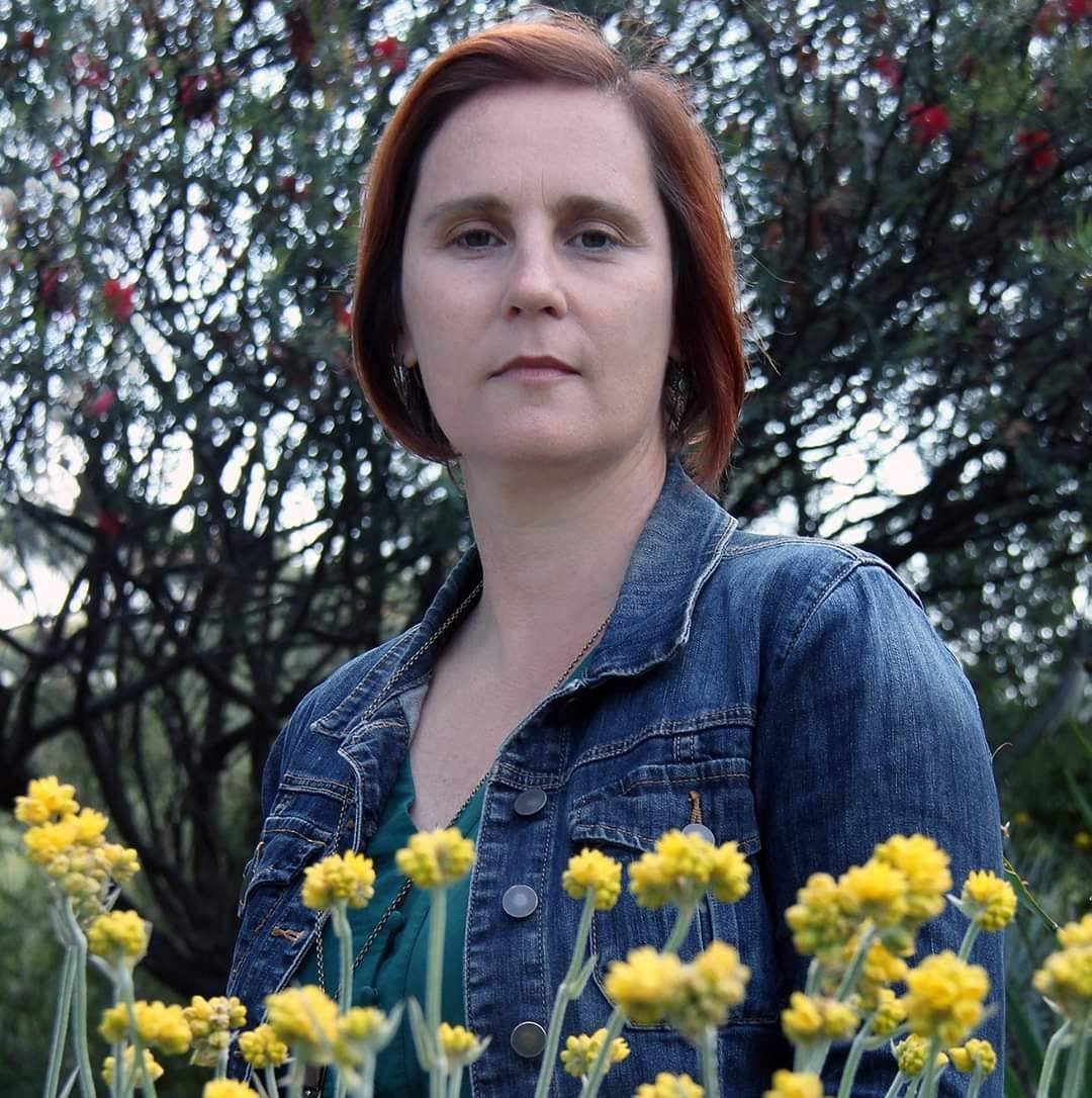 Natalie stands amongst flowers outside, wearing a denim jacket and looking at the camera.