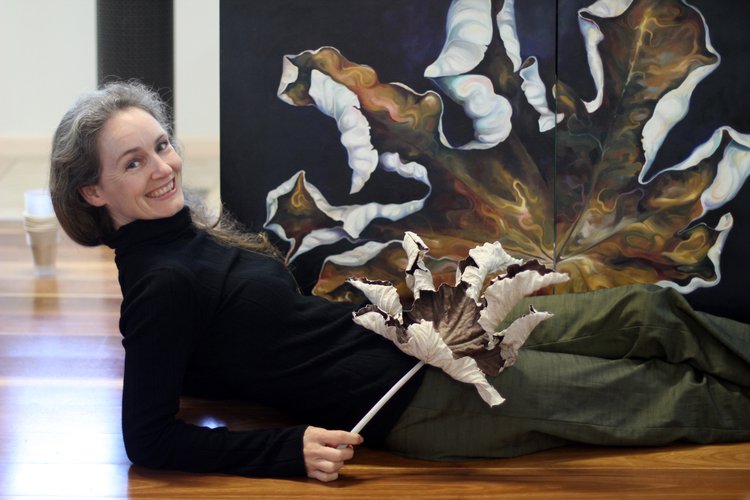 Tina Wilson lies in front of a painting of a dry leaf, while holding a large dry leaf.