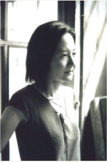 A black and white photo of Yoko who stands in the light coming from a window.