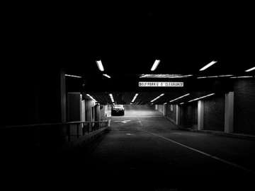 A dark, black and white image under a bridge. A car can be seen driving towards the camera.
