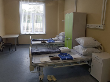 A dimly lit hospital room with light yellow walls and two beds. It's empty, but pyjamas are folded on each single bed as if patients are coming soon.